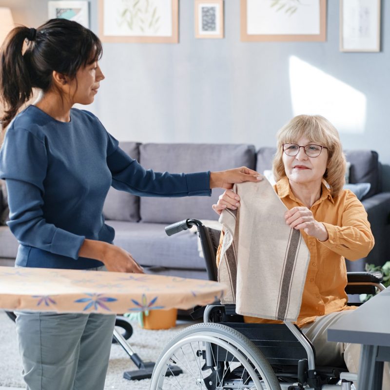 Housekeeper helping to iron the clothes to senior woman who using wheelchair at home